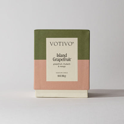 The Votivo Island Grapefruit 10 oz. Signature Candle, crafted with soy blend wax and featuring notes of grapefruit, rhubarb, and mango, comes boxed in green, pink, and cream color blocks against a light background.