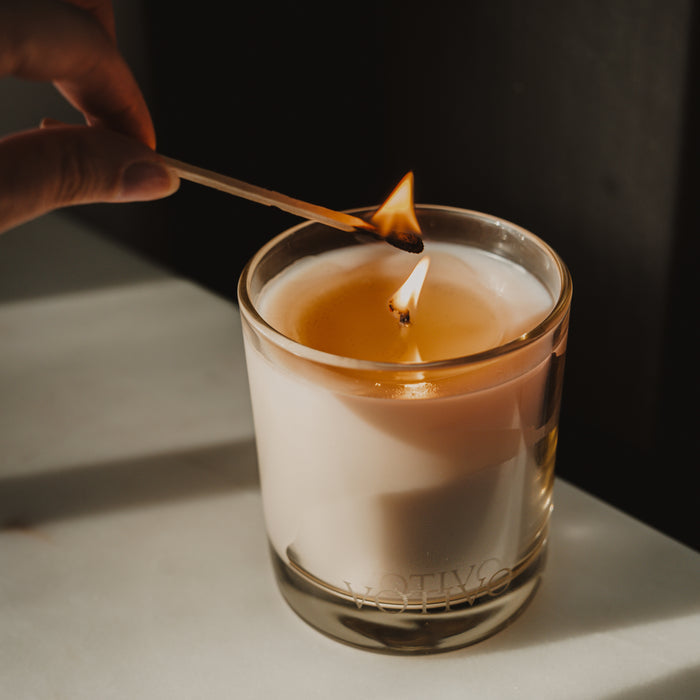A hand lights a Votivo Island Grapefruit 10 oz. Signature Candle in a glass jar with a matchstick. Made from soy blend wax, the candle sits on a light surface as its fresh fragrance begins to fill the air.