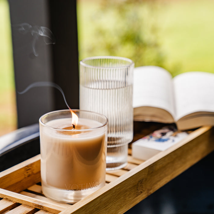 A lit Votivo Island Grapefruit 10 oz. Signature Candle, a glass of water, an open book, and a box of matches rest on a wooden tray. The soy blend wax releases a subtle scent as blurred greenery in the background creates a cozy, calming atmosphere.