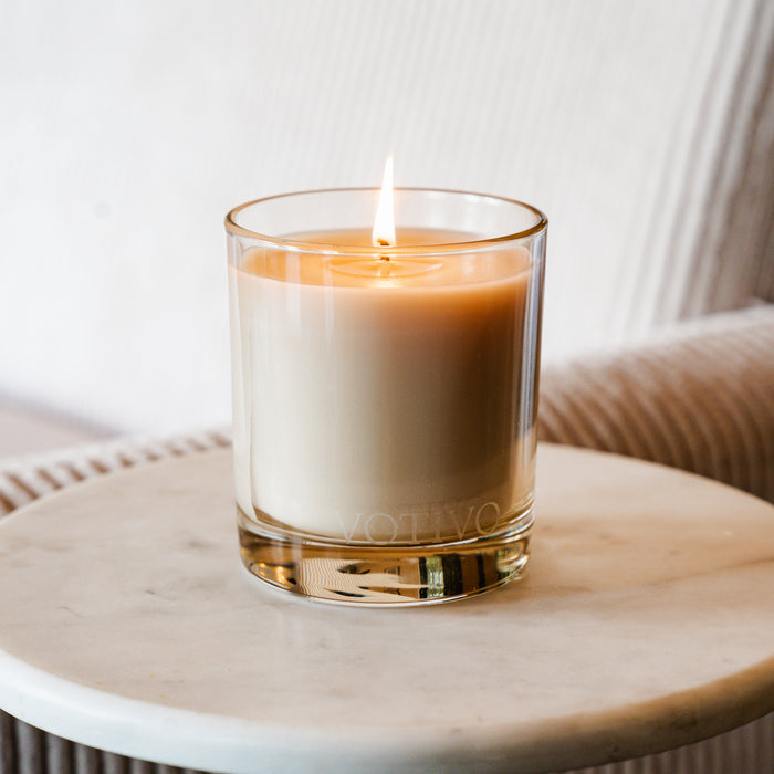 A lit Votivo Island Grapefruit 10 oz. Signature Candle made with soy blend wax sits in a clear glass container on a white marble surface, with a blurred light-colored couch and its fresh grapefruit scent in the background.