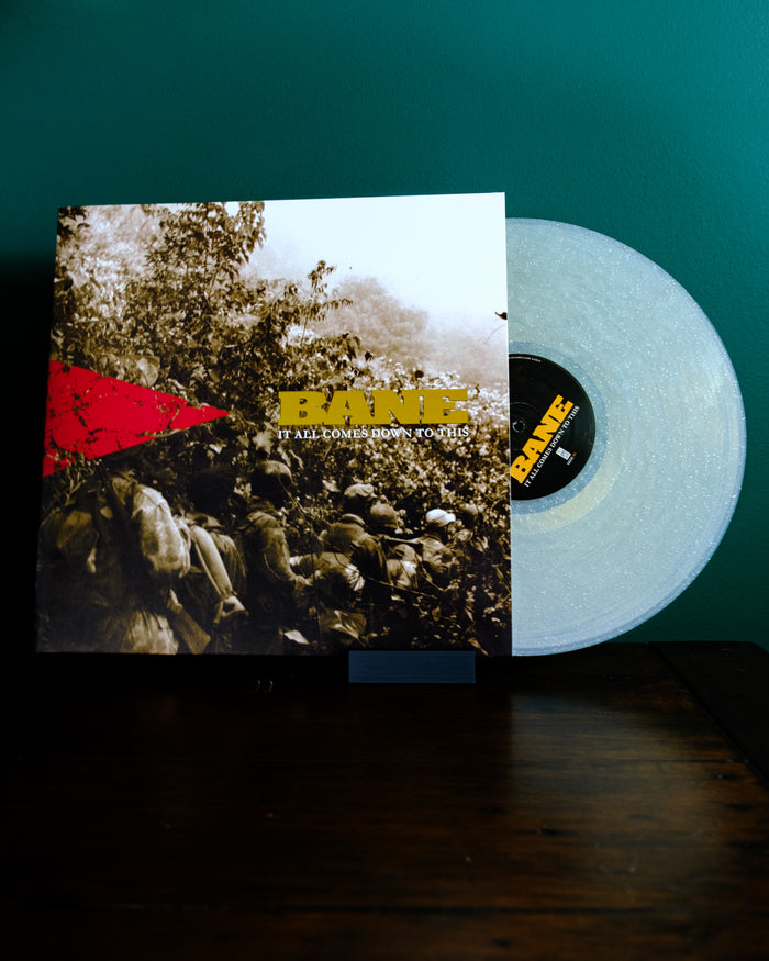 A translucent white vinyl record with a natural sparkle, by Bane, slides from an "It All Comes Down To This" cover featuring a sepia forest photo and red triangle design, set against a green wall.