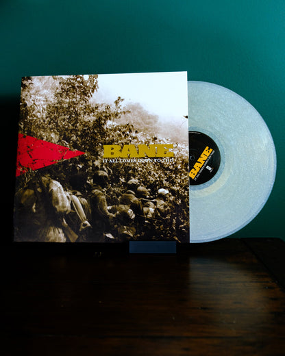 A translucent white vinyl record with a natural sparkle, by Bane, slides from an "It All Comes Down To This" cover featuring a sepia forest photo and red triangle design, set against a green wall.