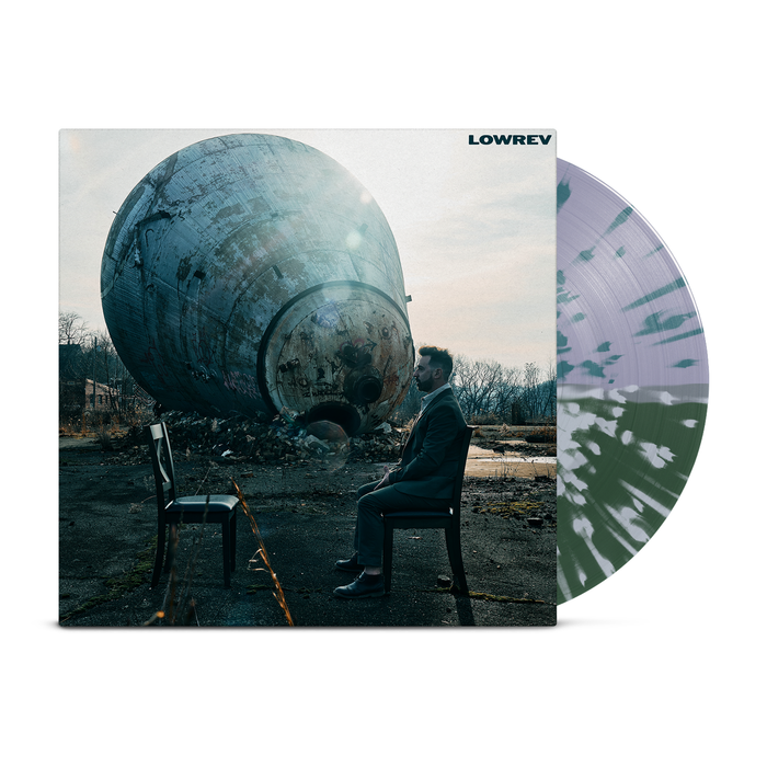 A man in a suit sits on a chair facing an empty one outdoors, with a weathered sphere behind him. The Lowrev album "It’s Hard to Lie to Strangers" is featured, including a signed postcard and green-purple streaked white vinyl.
