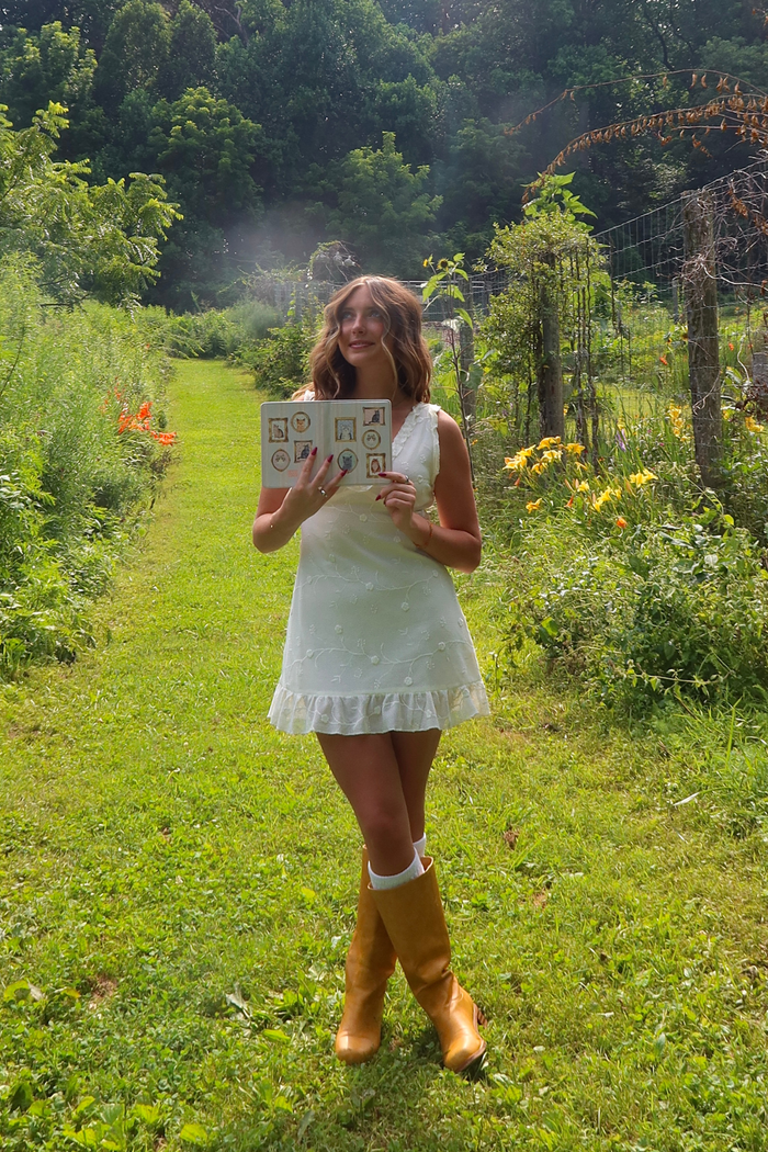A woman stands on a grassy path in a lush garden, wearing a white sleeveless dress and mustard yellow rain boots while holding a small printed card.