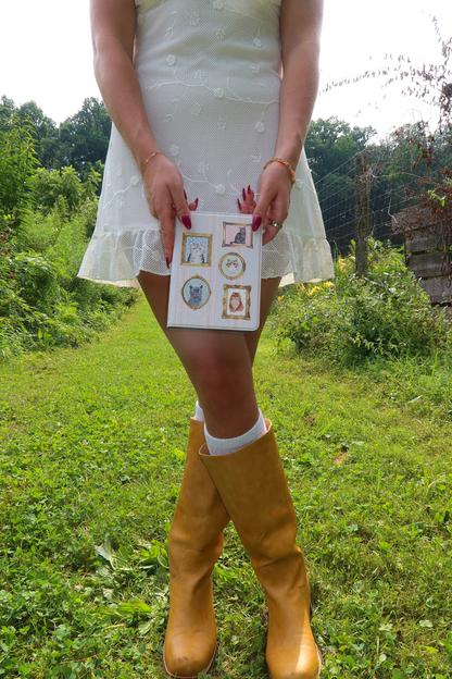 A person outdoors in a white dress and mustard yellow rain boots holds a small white card with several small framed illustrations.
