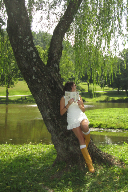 A woman in a white dress and yellow rain boots leans against a large tree next to a pond, holding a book.
