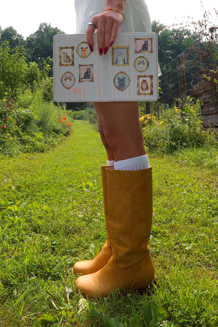 A person standing outdoors on a grassy path wearing tall mustard yellow boots and white socks, holding a white notebook decorated with small illustrated cat portraits.