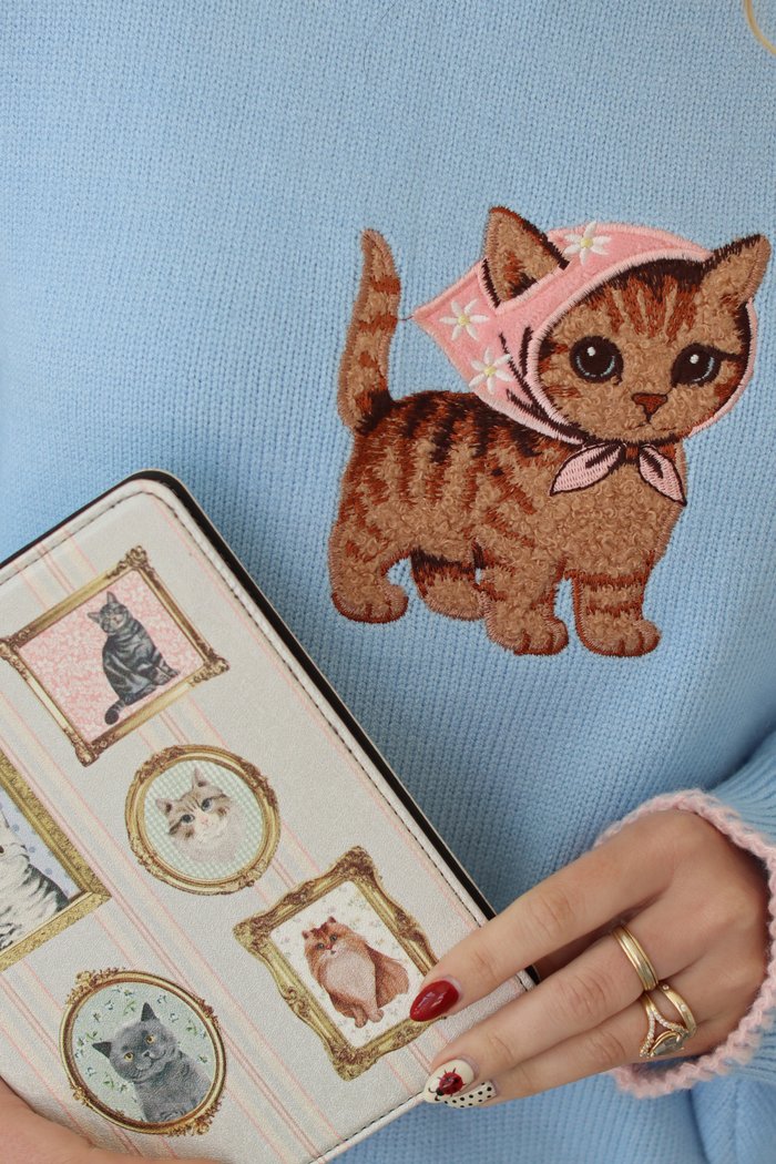 A close-up of a light blue knit sweater featuring a large embroidered brown tabby kitten wearing a pink bonnet. A hand with red nail polish and gold rings holds a decorative notebook with vintage-style cat illustrations.