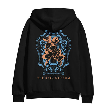 The back of the hoodie is a blue and orange colored crest design with THE RAIN MUSEUM written in orange text below it.