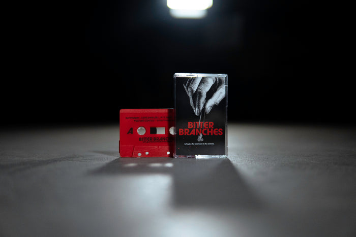 A red Let's Give The Land Back to the Animals cassette tape and case by Bitter Branches, featuring a hand image, rest on a gray surface under bright light against a dark backdrop—ideal for your album presale.