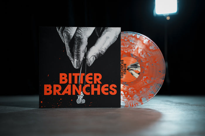 A Bitter Branches "Let's Give The Land Back to the Animals" orange and white marbled vinyl stands next to its cover, which features puppet strings held by a hand and “BITTER BRANCHES” in bold orange on black.