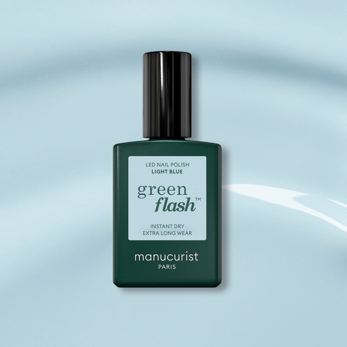 A bottle of Manucurist Light Blue Green Flash gel polish, with a dark green cap, sits on a soft blue background. The label highlights instant dry and extra long wear for this pastel sky blue shade.
