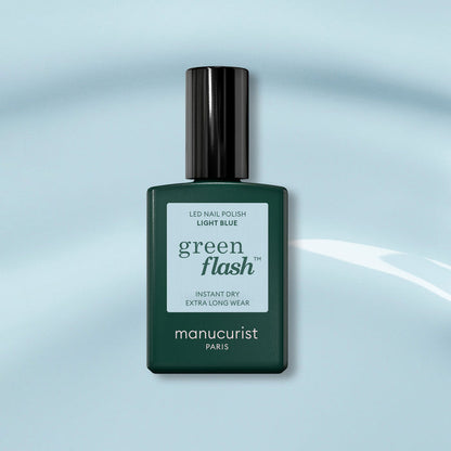 A bottle of Manucurist Light Blue Green Flash gel polish, with a dark green cap, sits on a soft blue background. The label highlights instant dry and extra long wear for this pastel sky blue shade.