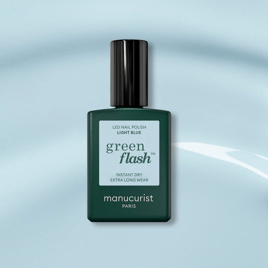 A bottle of Manucurist Light Blue Green Flash gel polish, with a dark green cap, sits on a soft blue background. The label highlights instant dry and extra long wear for this pastel sky blue shade.