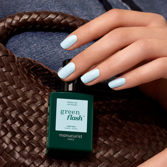 A hand with sky blue gel polish holds a bottle of Manucurist Light Blue polish, with a woven brown bag and denim fabric in the background.