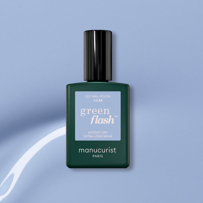 A bottle of Manucurist Lilas Green Flash gel polish, featuring a dark green base, black cap, and light purple label, is set against a light blue background with a white curved streak.