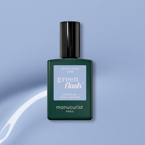 A bottle of Manucurist Lilas Green Flash gel polish, featuring a dark green base, black cap, and light purple label, is set against a light blue background with a white curved streak.