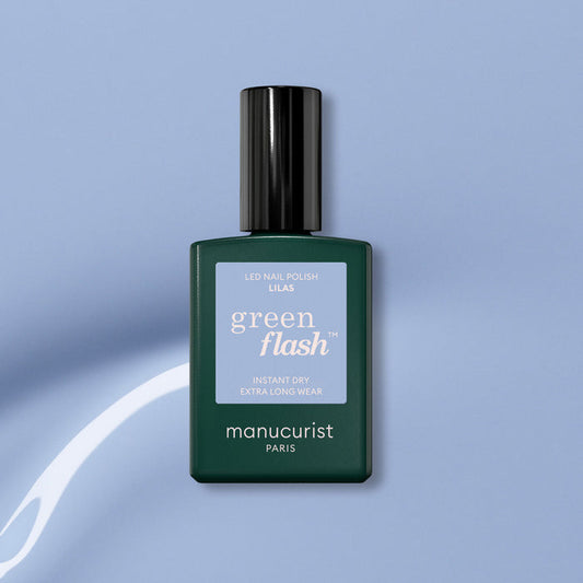 A bottle of Manucurist Lilas Green Flash gel polish, featuring a dark green base, black cap, and light purple label, is set against a light blue background with a white curved streak.