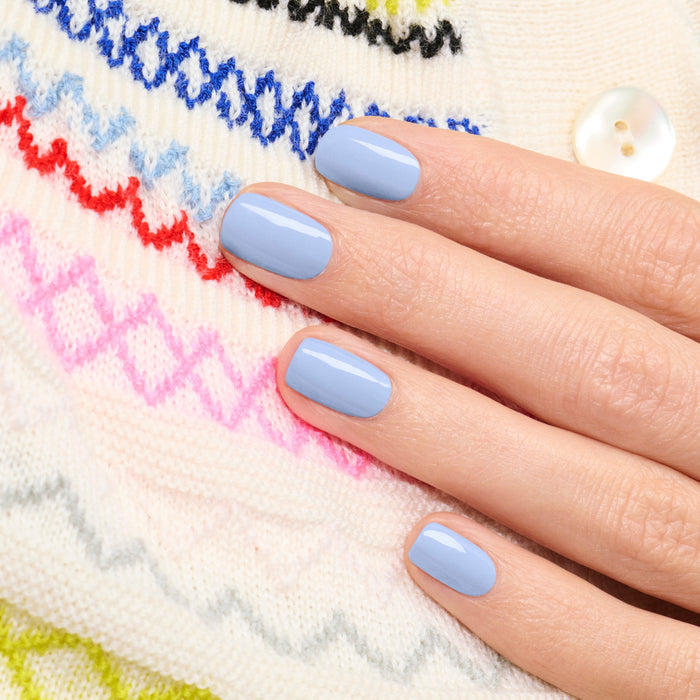 A hand with neatly manicured nails painted in Manucurist's Lilas pastel blue gel polish rests on a white sweater featuring colorful zigzag patterns and a white button.