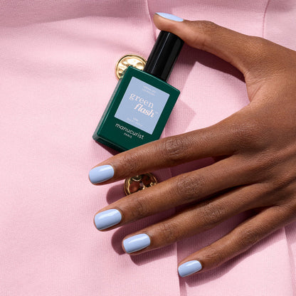 A hand with pastel blue gel polish holds a green bottle of Manucurist Lilas gel polish against a pale pink fabric background with a gold button.