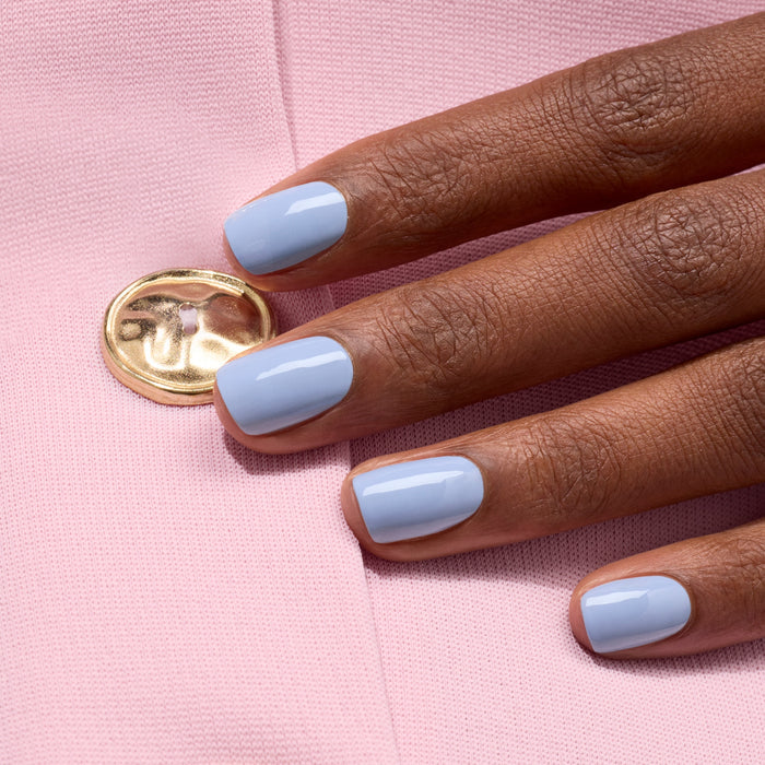 A hand with short, neat nails painted in Manucurist's glossy pastel blue Lilas gel polish rests on light pink fabric featuring a gold button.
