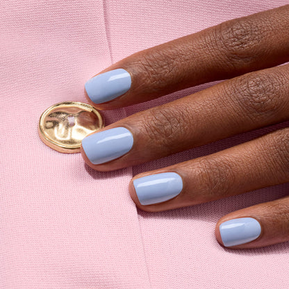 A hand with short, neat nails painted in Manucurist's glossy pastel blue Lilas gel polish rests on light pink fabric featuring a gold button.