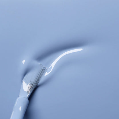 A white nail brush glides through Manucurist’s Lilas, a glossy pastel blue gel polish, leaving a textured curved streak on a smooth surface.