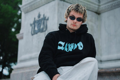 A young man wearing sunglasses, light pants, and a black Quicksand heavyweight pullover hoodie with a blue graphic and fleece-lined hood sits in front of a stone monument with engraved details and trees in the background.