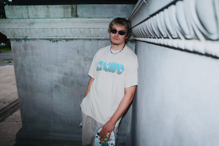 A young man wearing sunglasses and a light Quicksand garment-dyed t-shirt leans against a stone wall outdoors, holding a colorful skateboard.