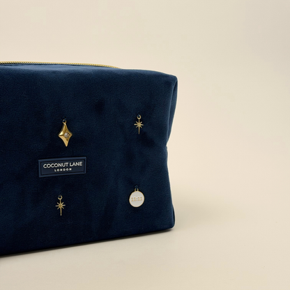 Stardust After Dark makeup bag. Dark navy blue velvet material with gold zip, and sewn-on gold celestial-themed charms including a sun, moon, star, diamond shape and 11:11 charm.