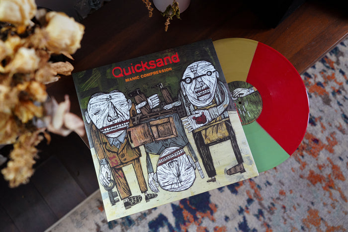 A Quicksand 30th Anniversary Deluxe Edition Manic Compression vinyl rests on a colorful rug. The split red, yellow, and green record matches the abstract, muted-toned album cover featuring distorted human figures.