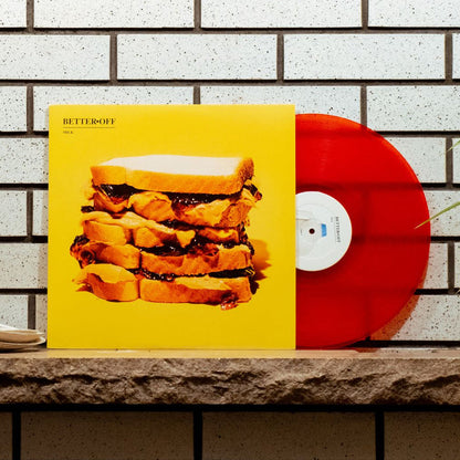 Yellow vinyl jacket with an image of a multiple layered sandwich. In the top left there is small text that says BETTER OFF, and below it there is smaller text that says MILK. Peeking out of the jacket is a red colored vinyl. Both are being leaned up on a subway tile wall.