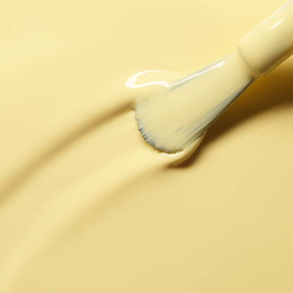 A close-up of a brush applying Manucurist's Mimosa, a creamy pastel yellow nail polish, onto a smooth yellow surface, adding subtle texture and gentle curves to the paint.