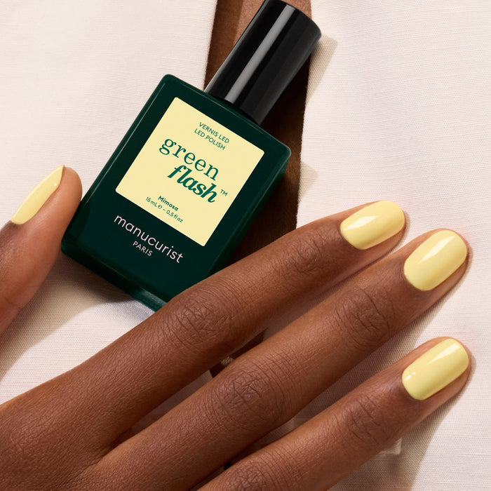A hand with neatly manicured pastel yellow gel polish in the shade Mimosa rests on a white fabric surface beside a bottle of Manucurist nail polish.