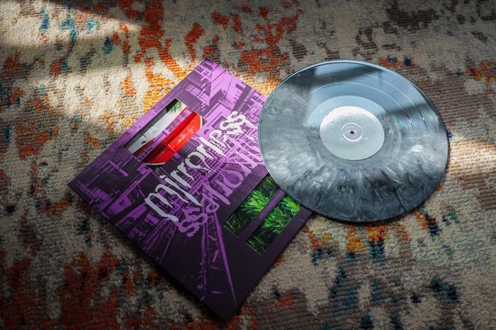 A limited edition marbled gray vinyl record by MIRRORLESS, partially out of its purple and green sleeve, rests on a patterned rug as sunlight streams in.
