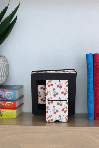 kindle with cherry pattern on a wooden surface with colorful books and a plant in the background.
