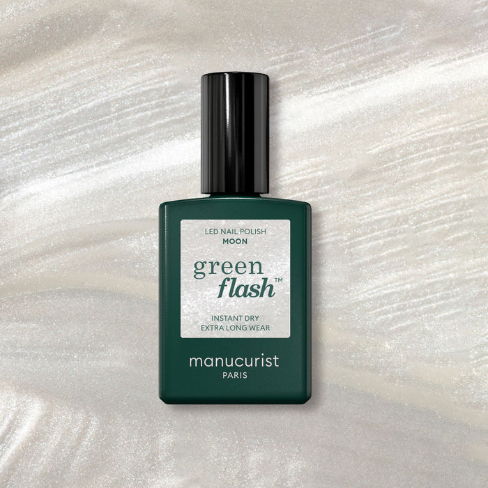 A bottle of Manucurist Moon gel polish rests on a shimmery marble background, its dark green design and white label highlighted by pearly white accents beneath.