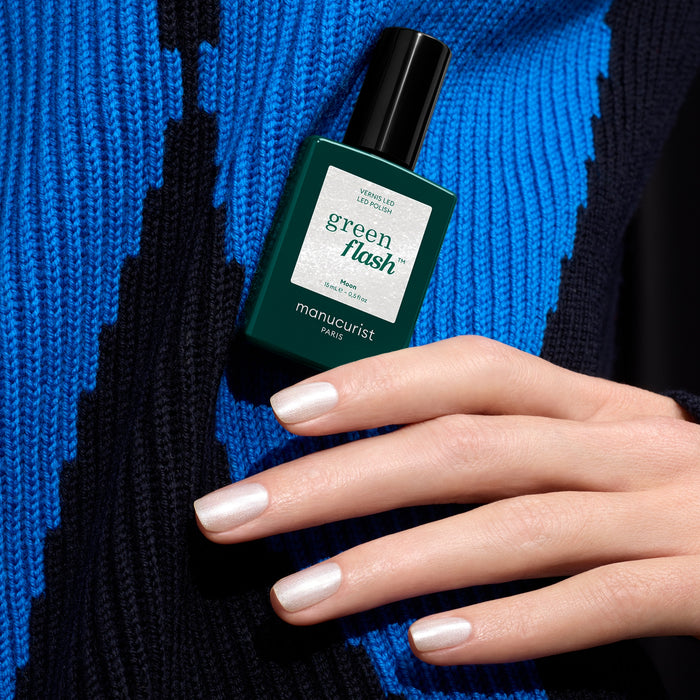 A hand with pearly white, shimmery nails holds a bottle of Manucurist Moon polish against a blue and black patterned background.