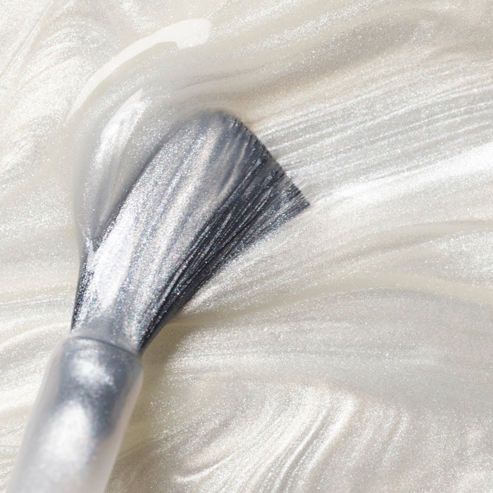 A close-up of a makeup brush spreading Manucurist's Moon, a shimmering pearly white gel polish, on a smooth surface to create soft, metallic streaks.