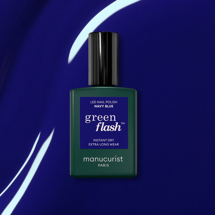 A bottle of Manucurist Navy Blue gel polish sits on a shiny deep blue surface with white curved light reflections.