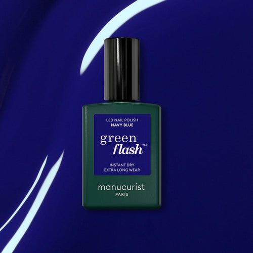 A bottle of Manucurist Navy Blue gel polish sits on a shiny deep blue surface with white curved light reflections.