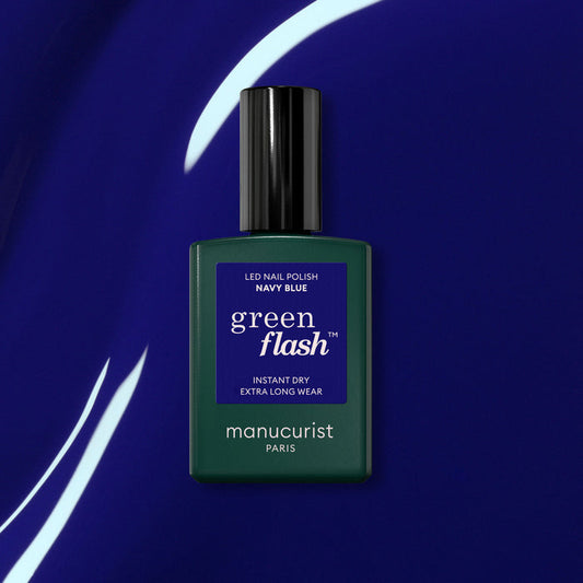 A bottle of Manucurist Navy Blue gel polish sits on a shiny deep blue surface with white curved light reflections.