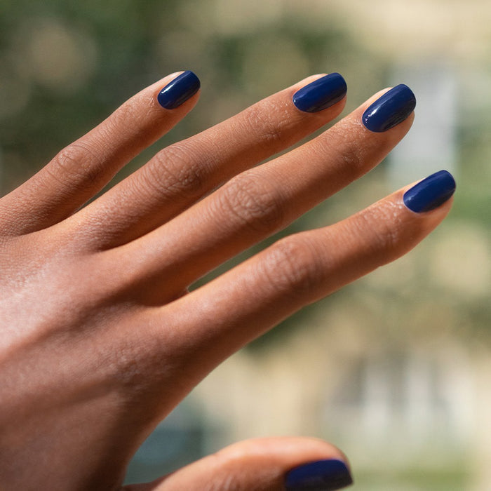 A hand with dark skin and neatly manicured nails is shown, each nail painted in Manucurist’s Navy Blue gel polish, against a softly blurred outdoor background.