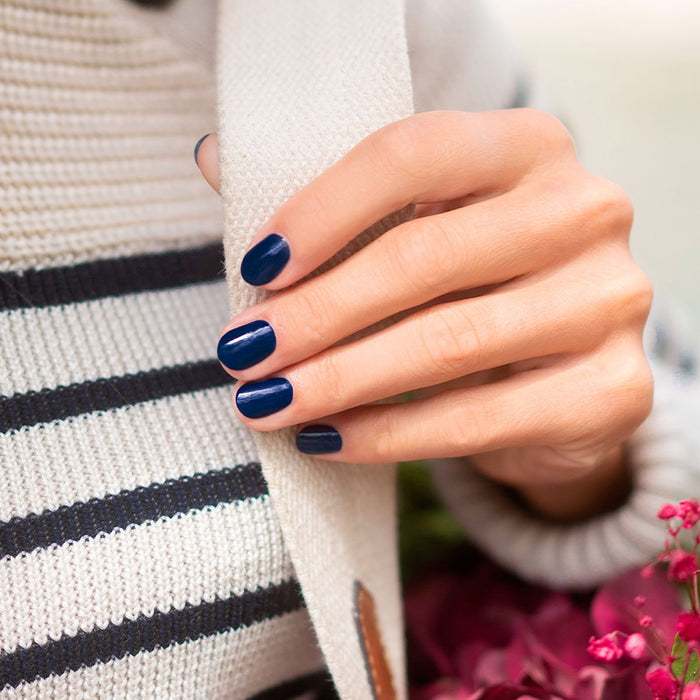A hand with neatly manicured nails painted in Manucurist Navy Blue gel polish rests on a cream knit scarf. The person wears a white and navy striped sweater, with pink flowers partly visible in the bottom right corner.