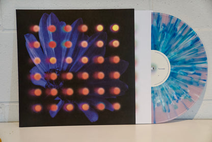 Anberlin’s Splatter LP, showcasing a pink and blue design, is partially pulled from its album cover with a dark flower and blurred orange dots—displayed on a white surface. Perfect for fans of Never Take Friendship Personal.