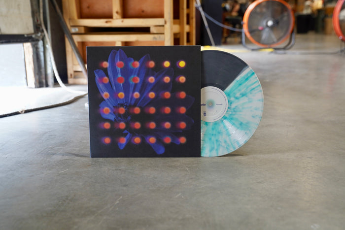 Anberlin’s Never Take Friendship Personal vinyl, in a blue and white marbled design, is half inside its sleeve with an abstract flower and red dots, standing upright on a concrete floor in an industrial setting.