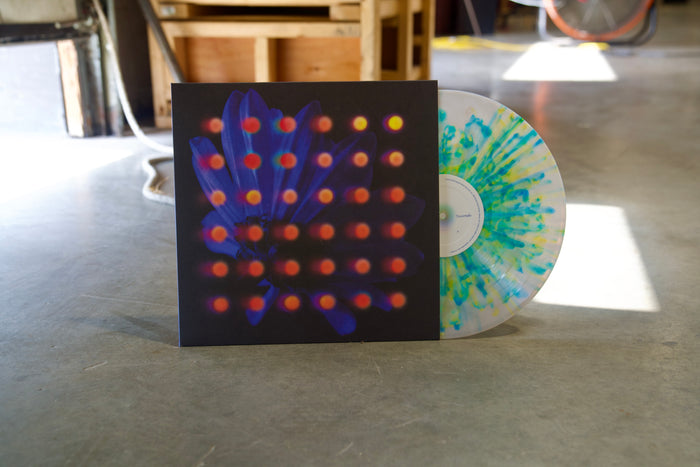 An Anberlin Lite Grey Marble LP with blue and yellow splatters leans against its abstract cover on a sunlit concrete floor—grab this presale vinyl before the album drops in 2025.
