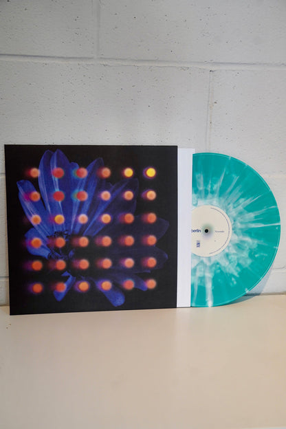 Anberlin's Nevertake Lite Teal LP presale vinyl features a turquoise and white marbled design, partially in a sleeve with abstract purple flower art, orange dots, and a black background against a white brick wall.