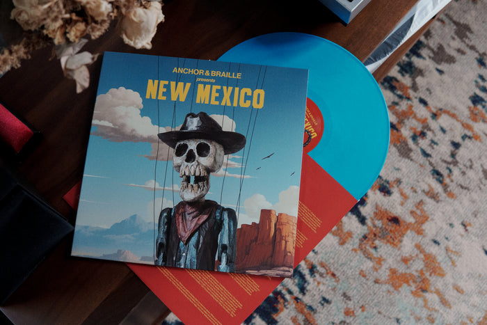 A blue vinyl LP and its cover lie on a table. The cover shows a skeleton in a cowboy hat and coat with "Anchor & Braille presents New Mexico" at the top, highlighting this striking Anchor & Braille vinyl album release.