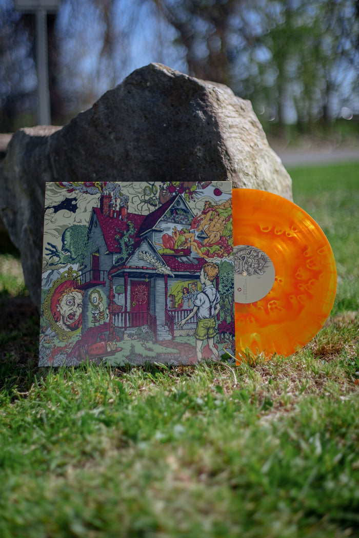 An orange cloud vinyl peeks out of a vinyl jacket with an illustration of a blue and red house with creepy figures and body parts coming out of it. The vinyl and jacket is propped up against a big rock outside in the grass.